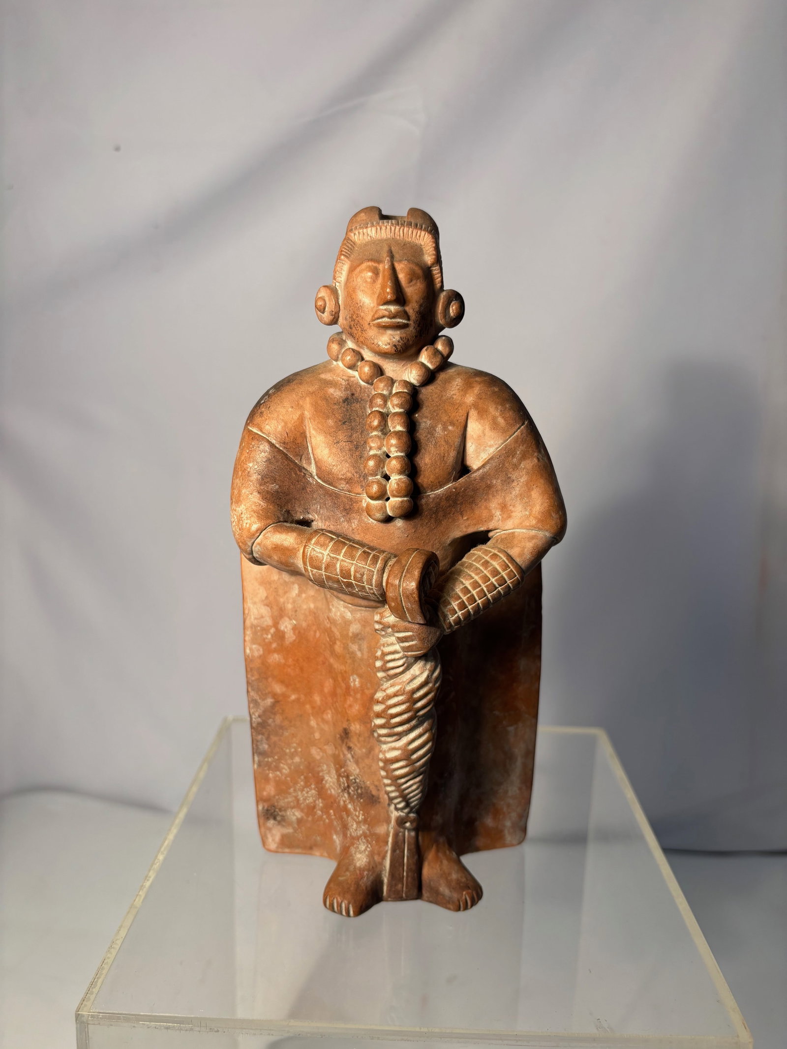 A Pre-Columbian Style Terracotta Standing Figure: Terracotta standing figure rendered in a Pre-Columbian style, depicting a stylized male figure with ear ornaments, necklace, and crossed arms. Red-brown surface with areas of patination and surface de