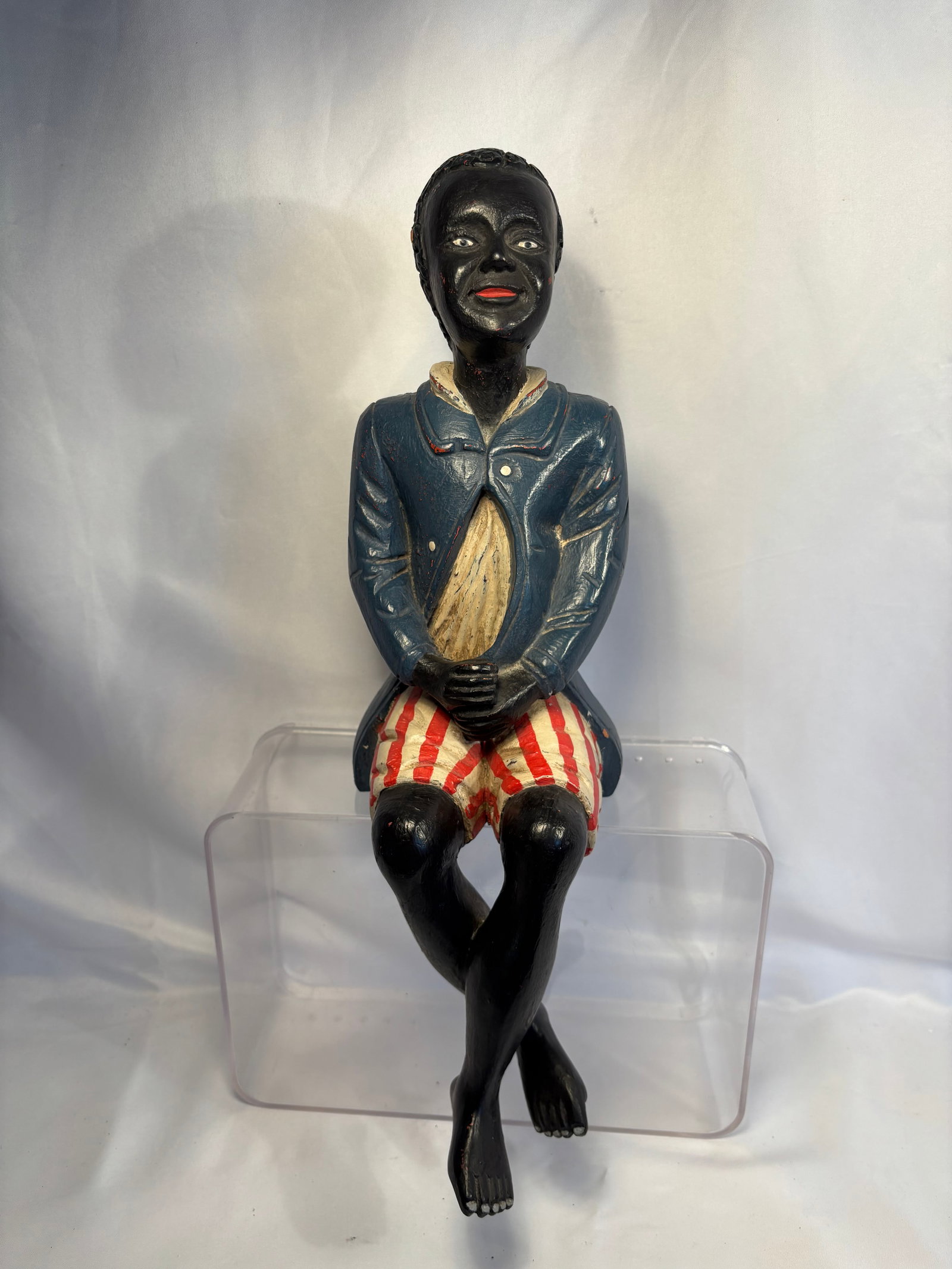 Antique American Folk Art Carved Wooden Black Americana Butler Figure: An antique American folk-art figure, carved and hand-painted as a seated Black butler or gentleman. The figure wears a blue jacket, cream-colored shirt, and red-and-white striped shorts, with folded h