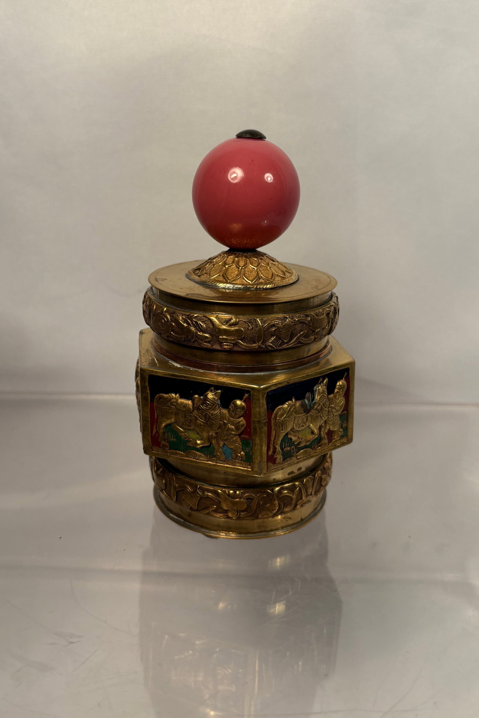 A Chinese Gilt-Brass Covered Container with Peking Glass Court-Style Finial: A decorative Chinese gilt-brass covered container of cylindrical and faceted form, richly embellished with raised foliate borders and figural panels depicting mounted and attendant figures against dar