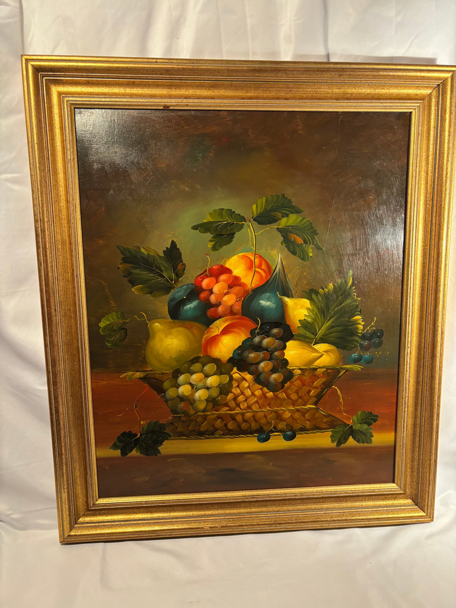 Framed Still Life with Fruit in a Woven Basket, Oil on Canvas: Oil on canvas still life depicting an abundant arrangement of grapes, peaches, pears, figs, and other fruit with broad green foliage, displayed in a woven basket upon a tabletop. Rendered in rich, sat