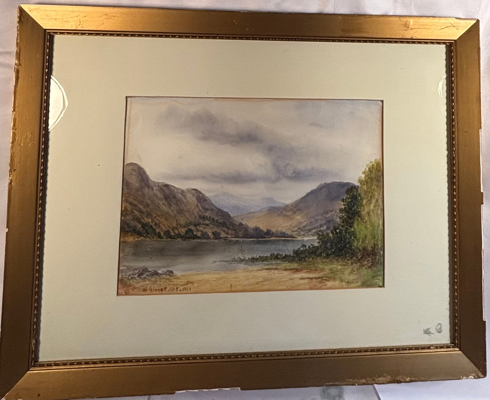 W. Glover, Mountain Lake Landscape, 1921, Watercolor: A watercolor on paper depicting a tranquil mountain lake landscape beneath a dramatic cloudy sky, with rugged hills rising along the shoreline and distant mountains visible through the valley. Execute