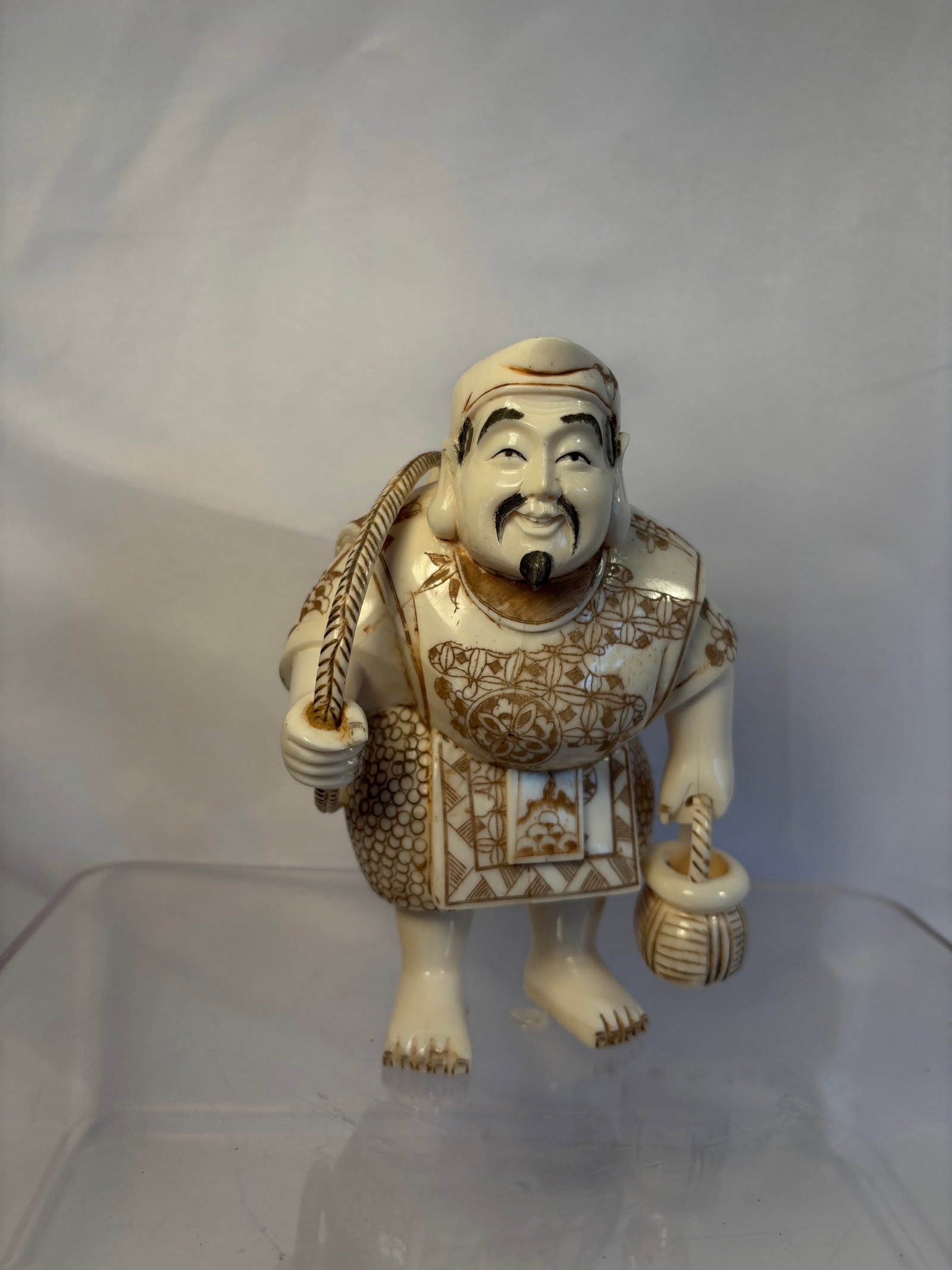 Japanese Carved Bone Okimono of Ebisu with Fish and Basket: A Japanese carved and engraved bone okimono depicting Ebisu, one of the Seven Lucky Gods and the patron of fishermen and good fortune. The smiling figure carries a large fish over his shoulder and hol