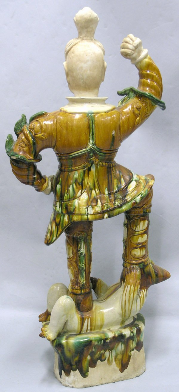 Chinese Tang-style sancai guardian: Chinese Tang-style sancai ceramic guardian figure, with one arm raised and standing on a humorous demon on a high plinth, the unglazed face with exaggerated express, the head and hands unglazed. 42.75