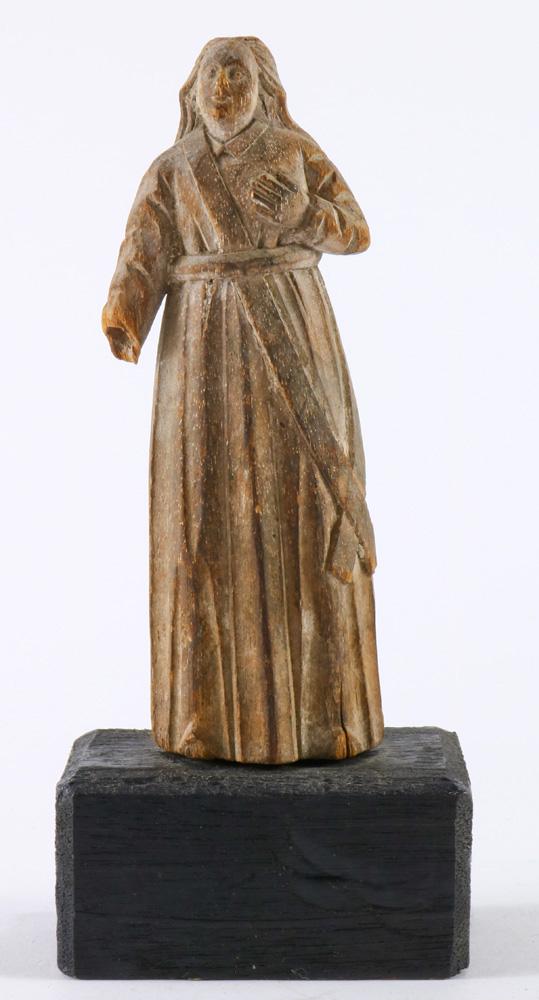A diminutive continental carved wood Santos on stand: A diminutive continental carved wood Santos on stand, late 18th / 19th century, the figure depicted with his left hand on his heart, and dressed in a long robe, rising on a later ebonized stand, overa