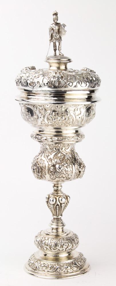 An Austro-Hungarian covered silver wine goblet: An Austro-Hungarian covered silver wine goblet, circa 1880, the repousse domical lid with a classical soldier finial, atop a waisted cup, the shoulder decorated with castle landscape reserves, rising 