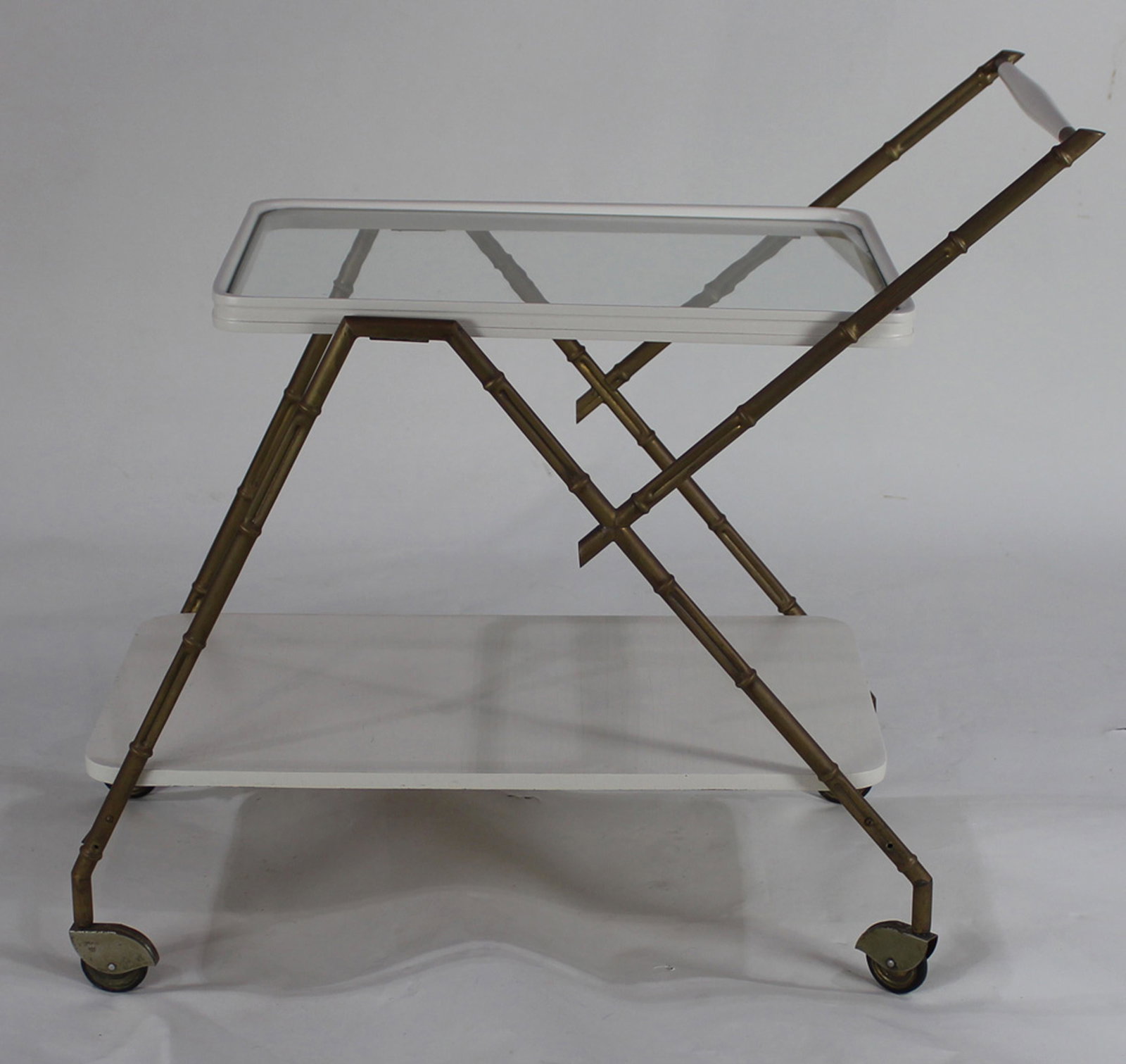 A Modern tea cart: A Modern tea cart, the tiered form having a glass top surrounded by the white apron, with a brass faux bamboo support conjoining the lower tier, and continuing to the casters, 28"h x 16"w x 29"d