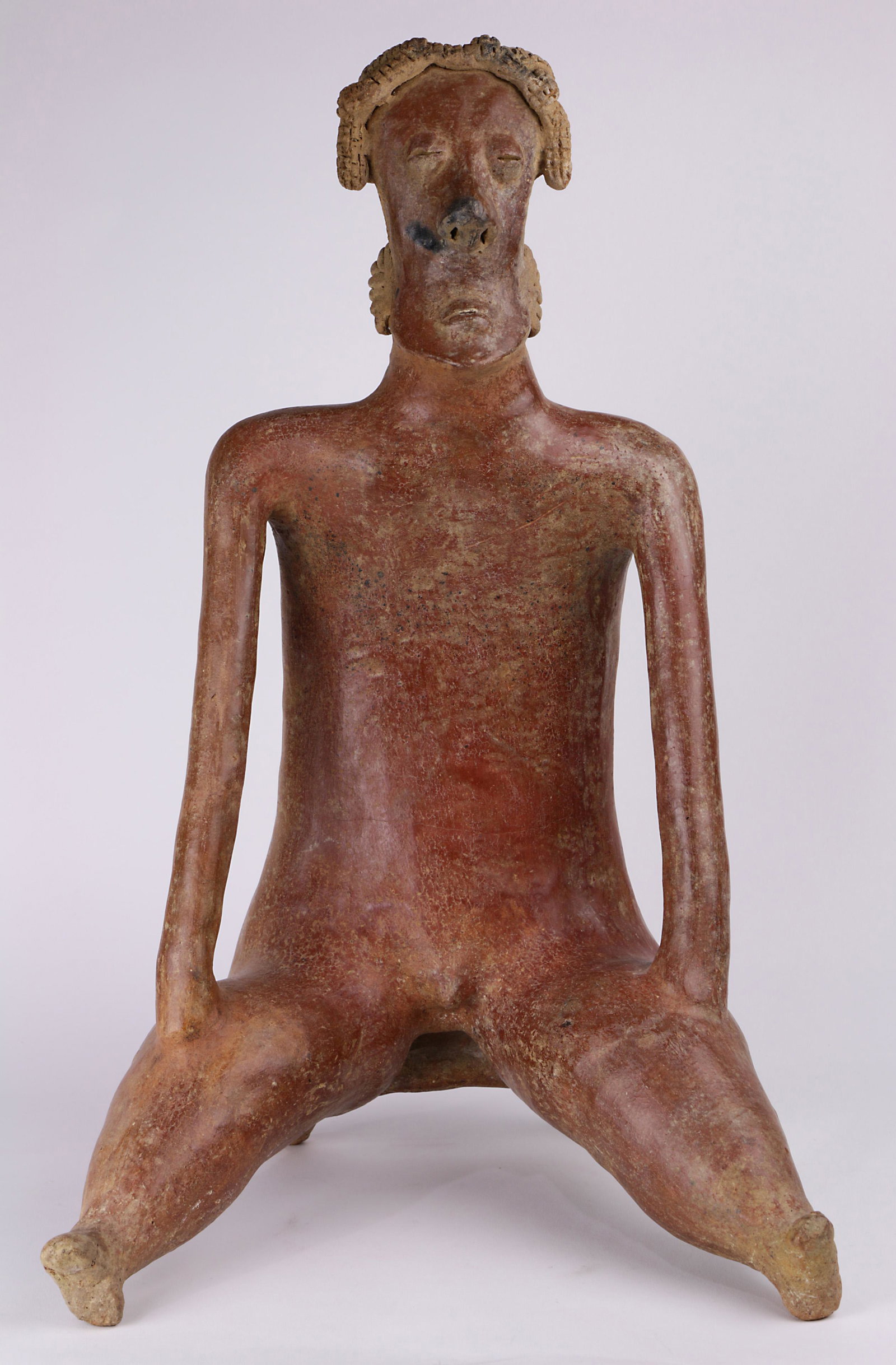 A Pre-Columbian Nayarit, West Mexico, seate male figure: A Pre-Columbian San Sebastian red regional type, Nayarit, West Mexico, seated male figure, 200BC-300AD, having an elaborate coiffure above stylized facial features, and depicted with hand on knees, 18