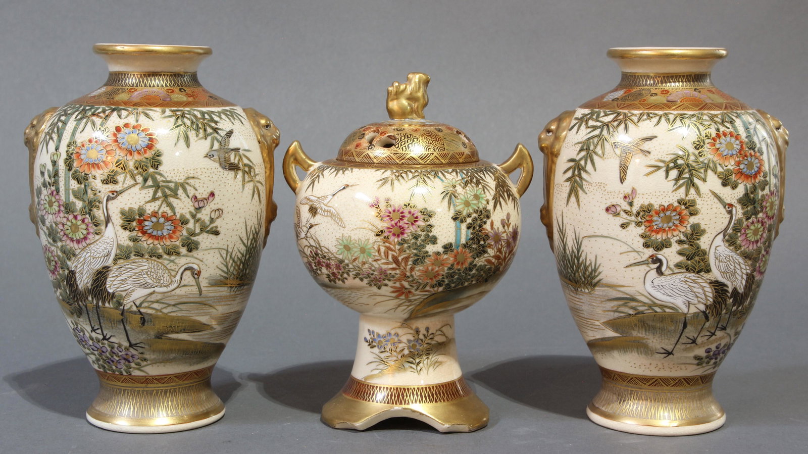 (lot of 3) Satsuma earthenware garniture: (lot of 3) Satsuma earthenware garniture, Kitamura Yaichiro, comprising a footed censer with shishi finial and a pair of urns with mock gilt shishi bust form handles, each enamel decorated with a gard