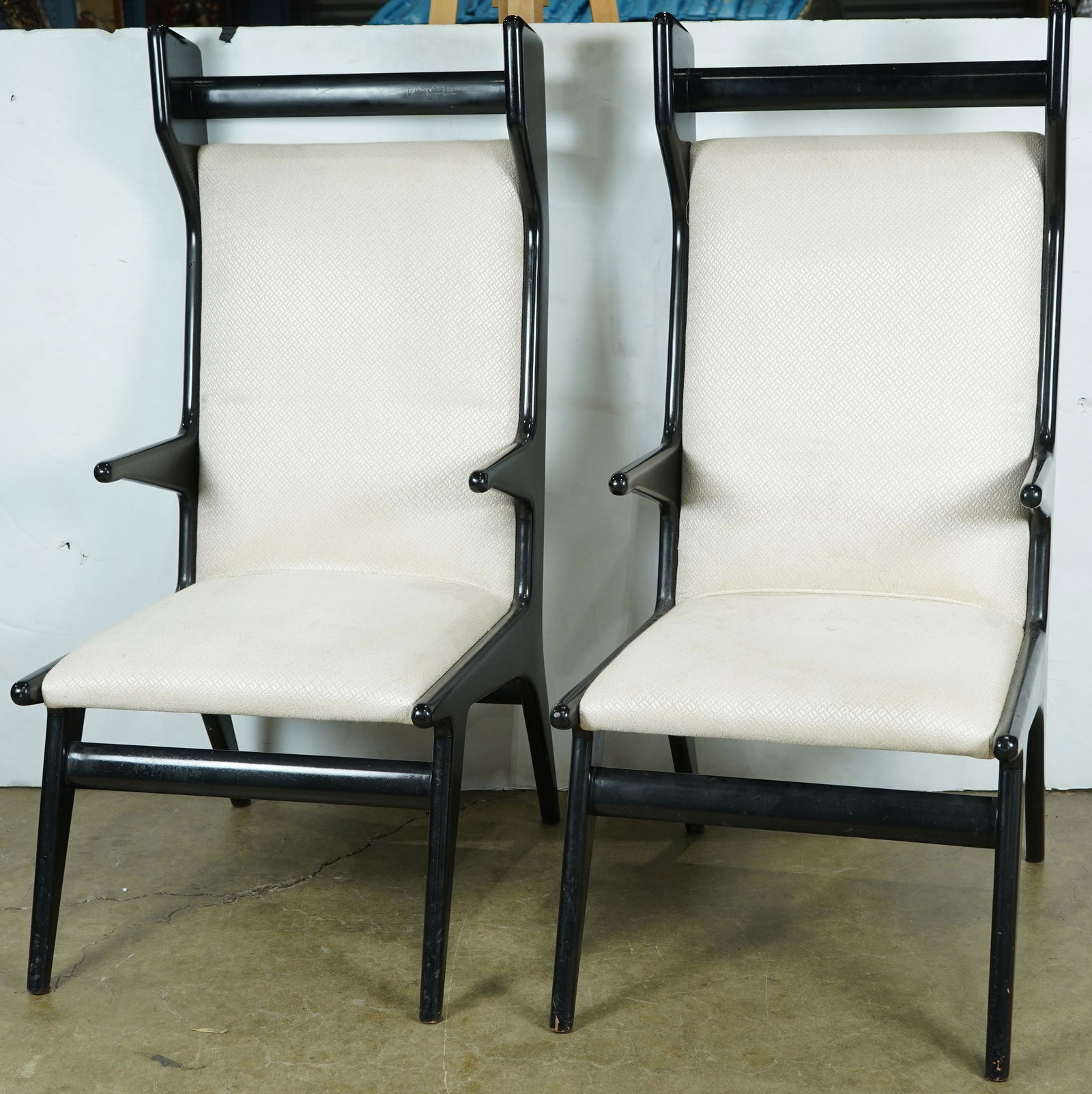 A pair of Italian Modern wing back chairs, in the: A pair of Italian Modern wing back chairs, in the manner of Gio Ponti, each having a black lacquer frame above cream upholstery and rising on tapered legs, 48"h