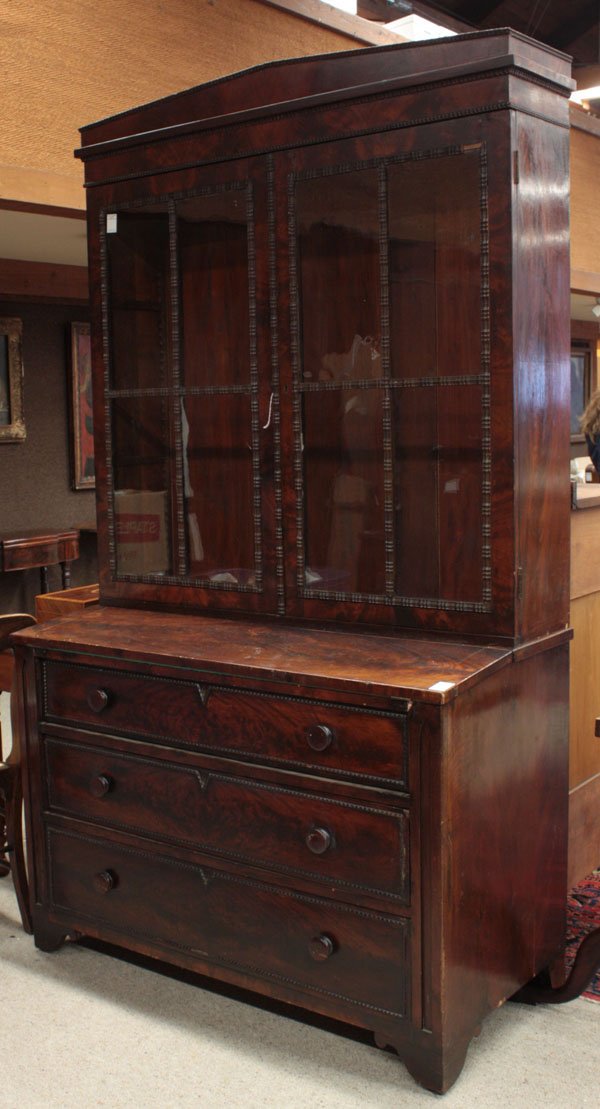 Victorian mahogany bookcase/chest: Two-part early Victorian flame mahogany bookcase with flip top baize writing surface, circa 1840, adorned in ribbon mouldings, pitched crest over double mullioned doors, upon base with three graduated