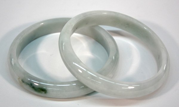 Chinese Carved Jade Bangle Bracelets: (lot of 2) Chinese green jade bangle bracelets, each with a rounded outer edge and with the semi-translucent pale green matrix flecked with white and darker green inclusions, 3.125''outer diam; 2.5''i