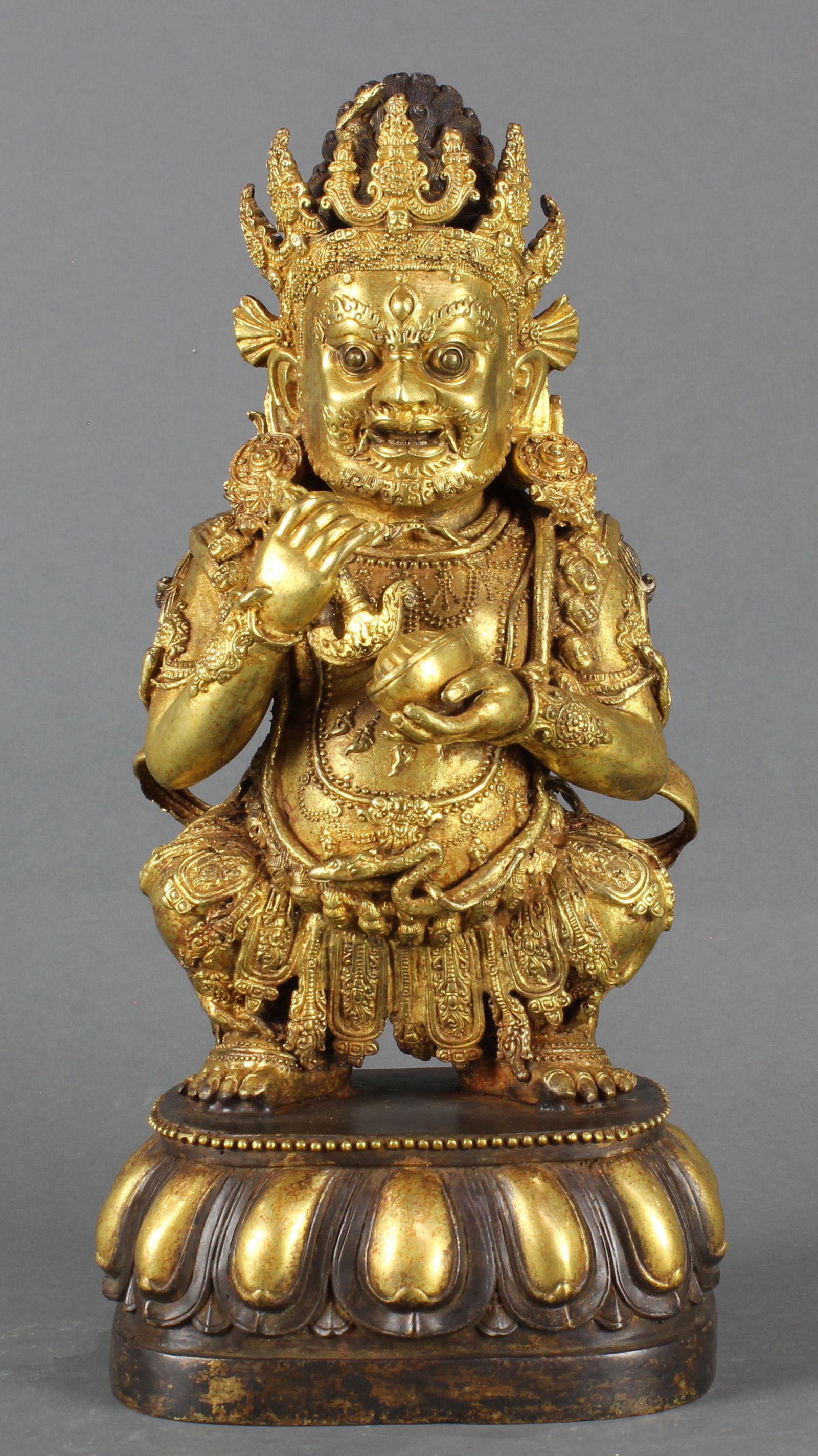 Gilt bronze figure of Mahakala: Gilt bronze figure of Mahakala, adorned with a necklace of heads and clutching a karttrka and kapala, the stern face flanked by elongated ears, wearing a crown, kneeling on a lotus thrown. 11.25"h.