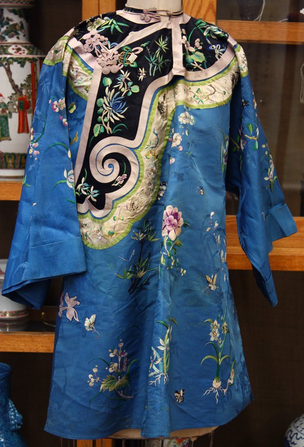 Chinese Woman's Blue Silk Robe: Chinese Han woman's silk robe, embroidered with butterflies and insects scattered amid various blossoming flowers upon a floral patterned blue damask ground, with similarly decorated trim (soiled and 