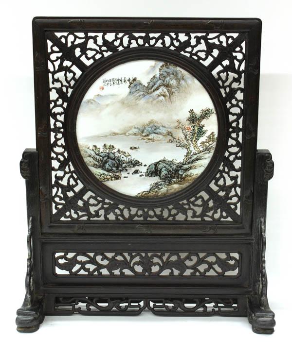 Enameled Chinese Porcelain Table Screen: Chinese hardstone table screen with a circular polychrome enameled porcelain plaque, painted in bright enamels with a river landscape bordered by stylized mountains, the upper left inscribed with a co