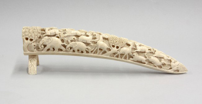 Ivory Tusk Carved with Animals: Carved ivory tusk, the contoured segment carved and undercut with a scene of a pack of lions chasing gazelles, giraffes and elephants, capped at the end with a budding rosette, the arched stand carved