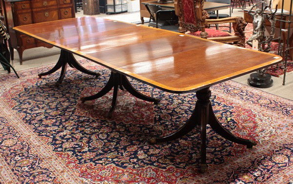 Regency style dining table: (lot of 4) Regency style dining table, the mahogany band inlaid top with three leaves and rising on a triple pedestal base having an acanthus carved standard with four outswept legs terminating on bra
