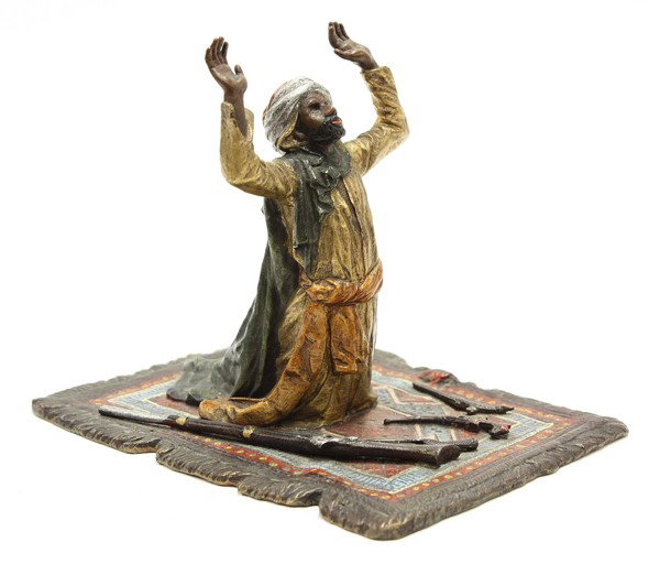 Austrian cold painted bronze: Cold painted bronze of a man in prayer, depicted with hands raised to the heavens, kneeling on a carpet and surrounded by his weapons, 4.5''h x 5''l