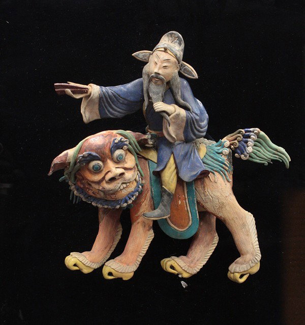Chinese Figural Architectural Ornament: Chinese glazed figural ceramic architectural ornament, fashioned as a Chinese sage stroking his long black beard while seated on the back of a humorous leonine beast with a blue and green curly mane a