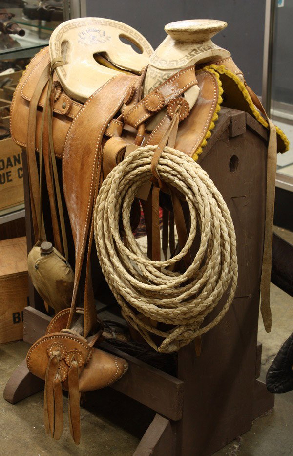 El Charro Mexican saddle: El Charro Mexican saddle with dee horn rigging, double rawhide covered skirt, together with a canteen, rawhide lasso, and a rifle scabbard, all on stand