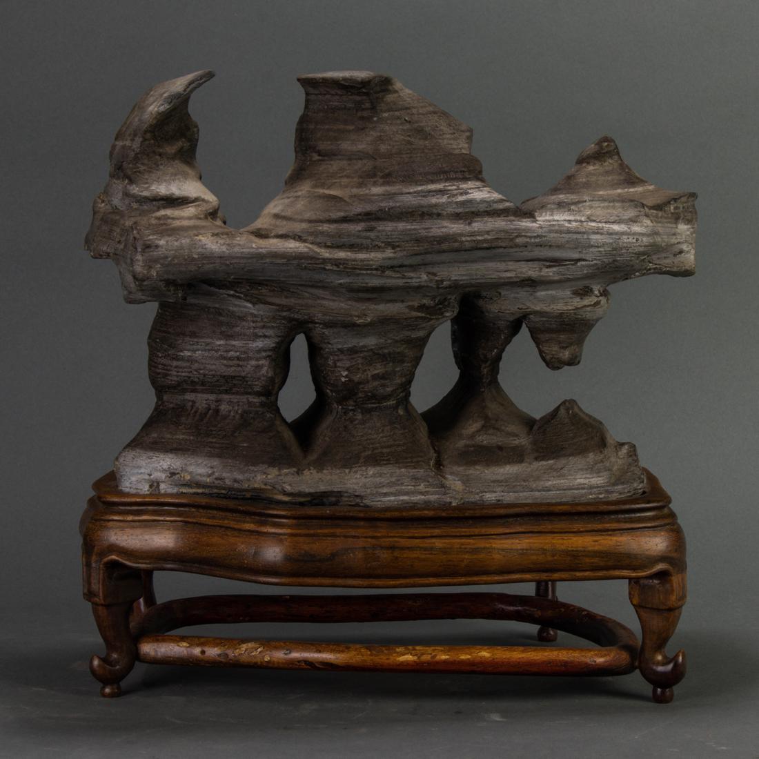 Chinese Qilian limestone scholar's rock: Chinese Qilian limestone scholar's rock, the striated gray stone displaying a series of three peaks above overhanging cliffs, Northern style wood stand, width 11.5".