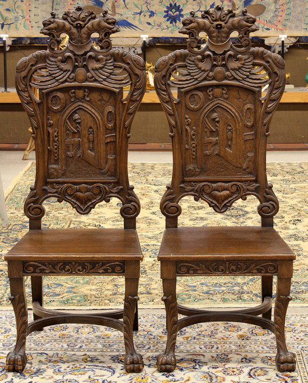 Continental style hall chairs: Pair of Continental style carved oak hall chairs circa 1900, each having a griffin carved crest continuing to the stiles and centering the panelled architectural back, and rising on carved legs termin