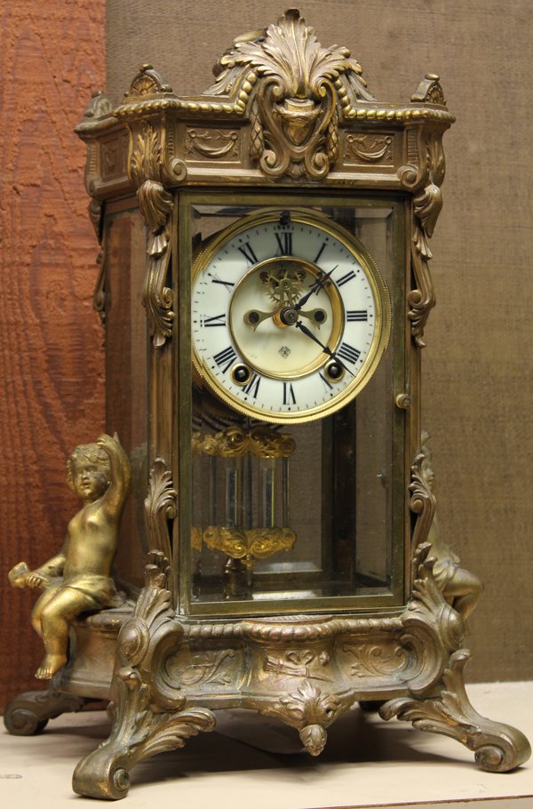 Ansonia mantle clock: Ansonia mantle clock, having a patinated metal case flanked with two seated putti, the bevelled glass windows opening to the Ansomia movement having an open escapement, and rising on inswept feet, 20'