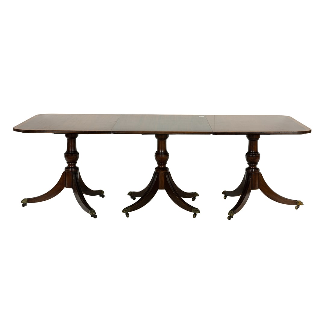 A Regency style mahogany pedestal dining table: A Regency style mahogany pedestal dining table, the crossbanded inlaid top with D-shaped ends, raised on three baluster standards, each issuing four outswept legs, ending in brass capped paw feet atop