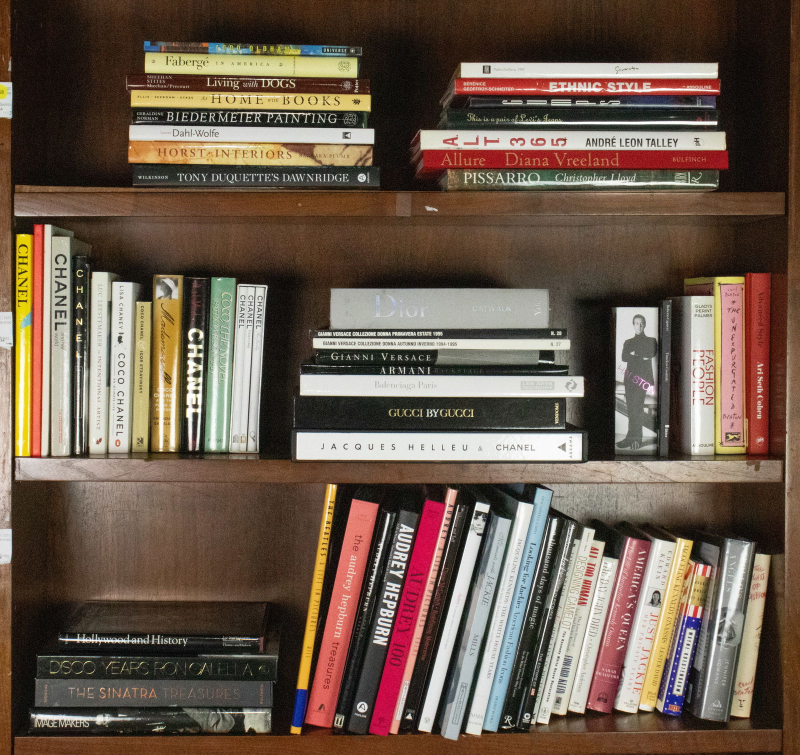 Three shelves of hardcover books pertaining to fashion brands and american fashion icons: Three shelves of hardcover books pertaining to household fashion brands and american fashion icons, including titles highlighting Chanel, Versace, Armani, Balenciaga, Gucci, Audrey Hepburn, and Nancy