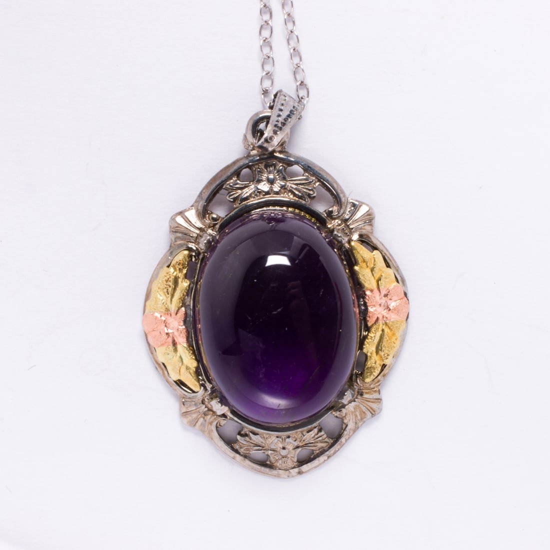 An amethyst and 14k tri-color gold pendant necklace: An amethyst and 14k tri-color gold pendant necklaceof floriform design, centering a oval amethyst cabochon measuring approximately 17.50 x 13.10 x 9.00 mm, suspending from silver cable-link chain;
