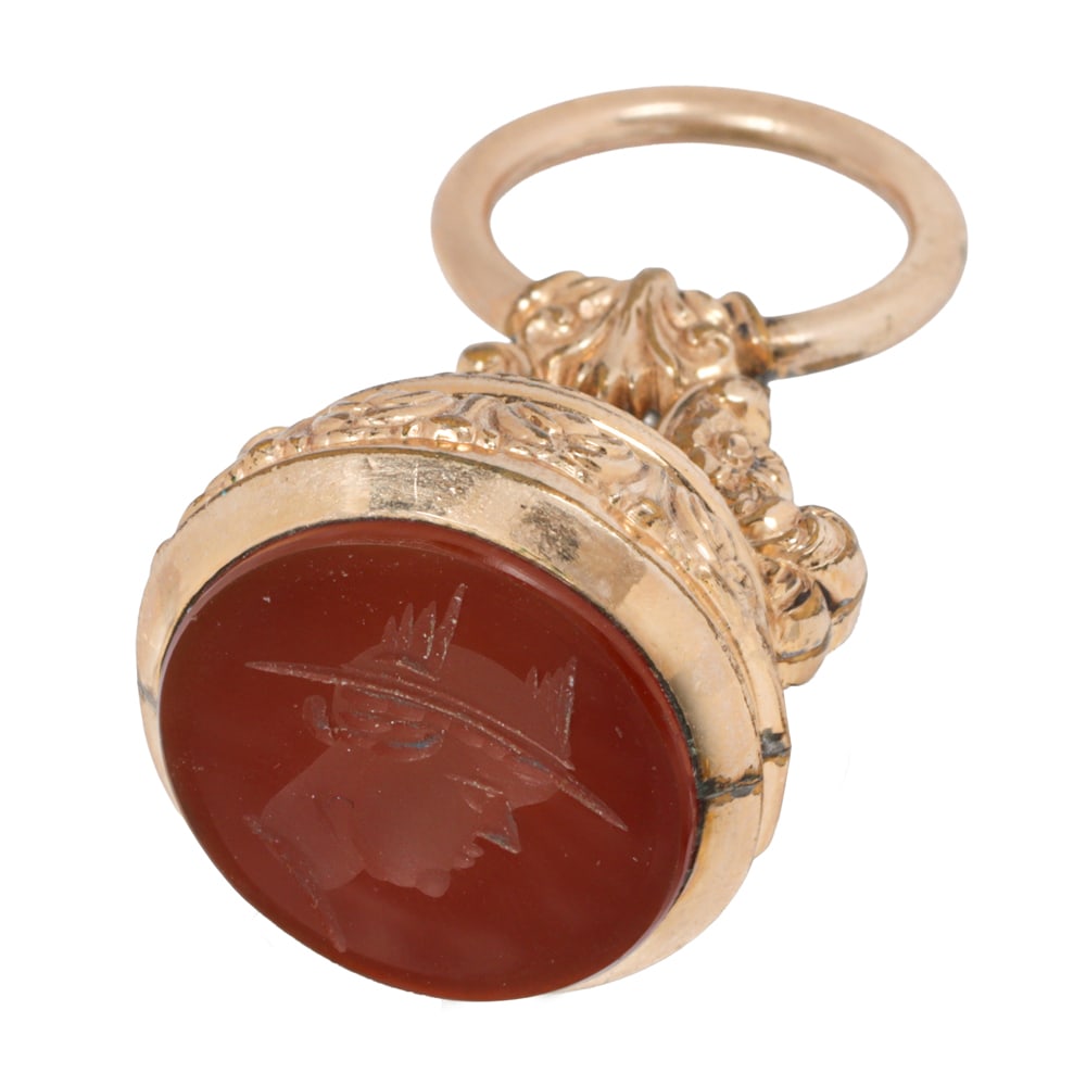 An antique 9k gold carnelian intaglio watch fob: An antique 9k gold carnelian intaglio watch fob Of hollow-form design, accented by embossed scrollwork and set with a carnelian intaglio; length: 1 3/4 in; 10.50 grams