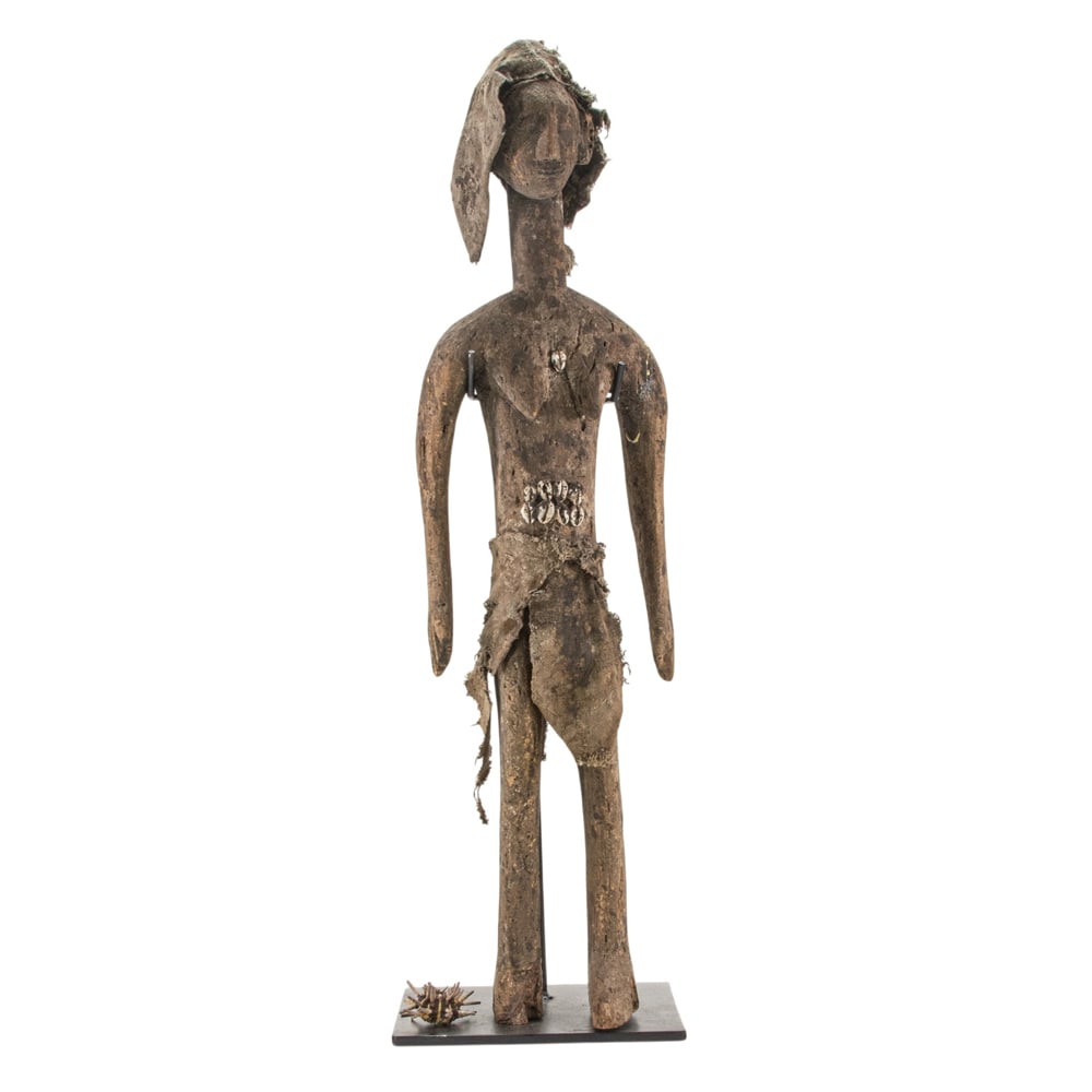 An African tribal figure: An African tribal figure, depicted standing wearing cloth cap and loincloth and further decorated with cowrie shells, raised on stand, 34"h