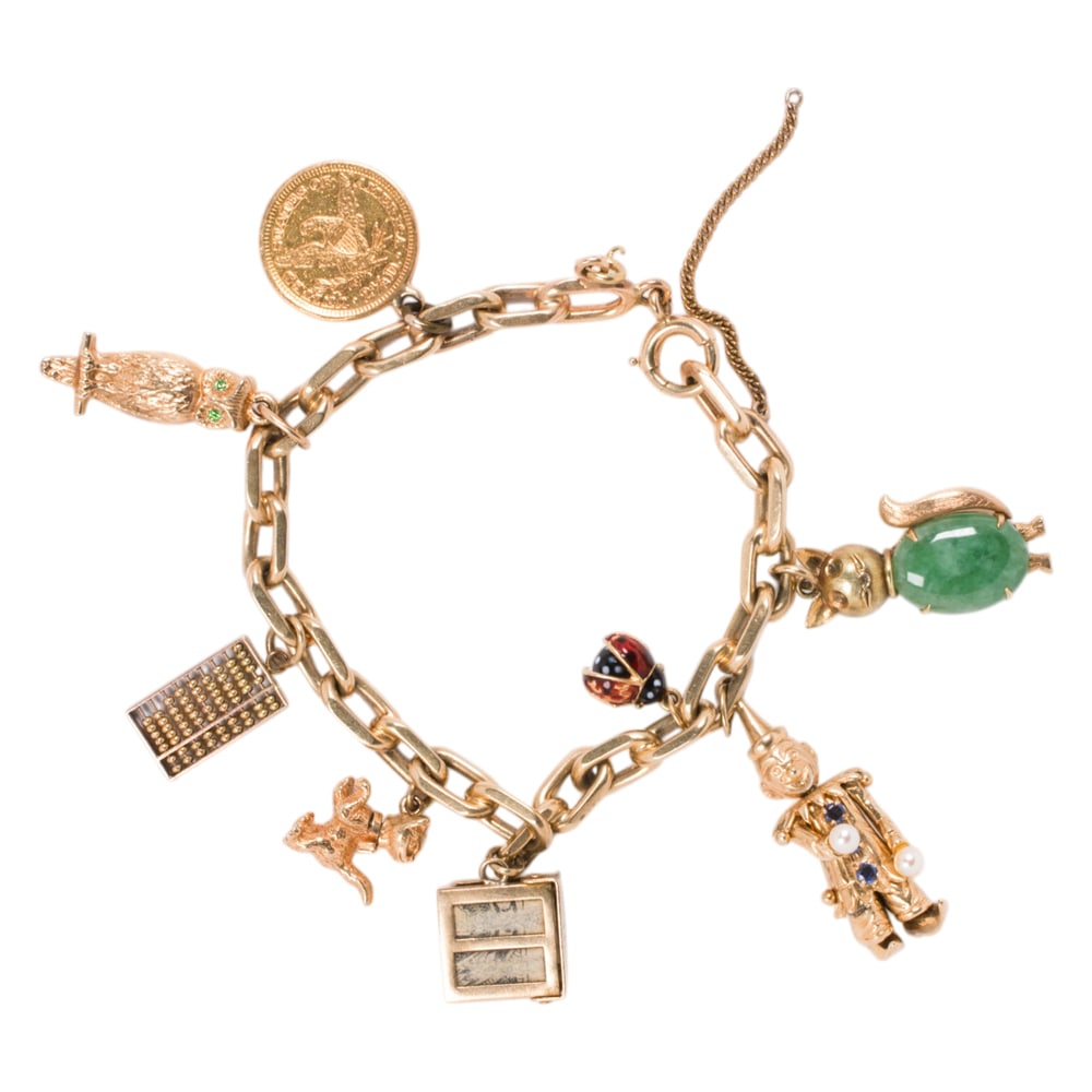 A 14k gold charm bracelet: A 14k gold charm bracelet featuring 8 charms consisting of a sapphire and cultured pearl set jester, a jade cabochon cat, emerald set owl, enamel lady bug, 2 1/2 USA gold coin, abacus, kitten, and mon