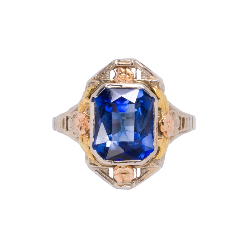 A synthetic sapphire and tri-color 14k gold ring: A synthetic sapphire and tri-color 14k gold ring Centering a fancy-cut synthetic sapphire weighing approximately 3.20 carats, within floriform tri-color gold setting; size 6 1/2; 3.45 grams