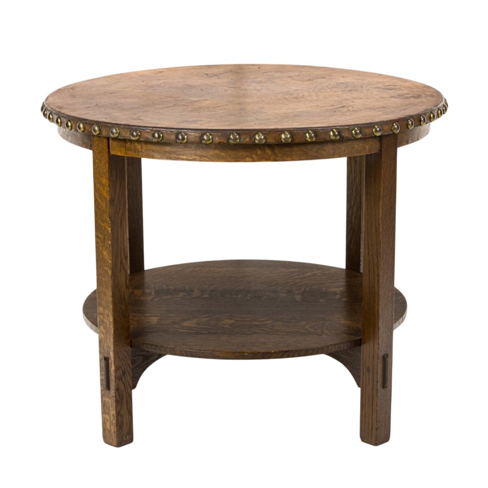 An Arts and Crafts Gustav Stickley leather top lamp table: An Arts and Crafts Gustav Stickley leather top lamp table, retaining original leather top with brass nail head trim, the lower tier with thru tenon supports, 29.5"h x 36'dia Provenance: Isak