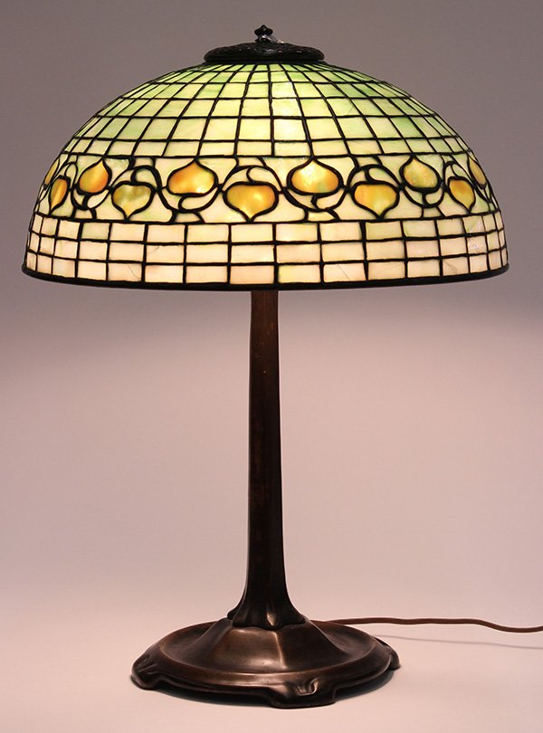 Tiffany Studios patinated bronze and leaded glass table: Tiffany Studios patinated bronze and leaded glass table lamp, the geometric decorated shade centered with a leaf and vine frieze, above the three socket cluster rising on a faceted footed base, shade 