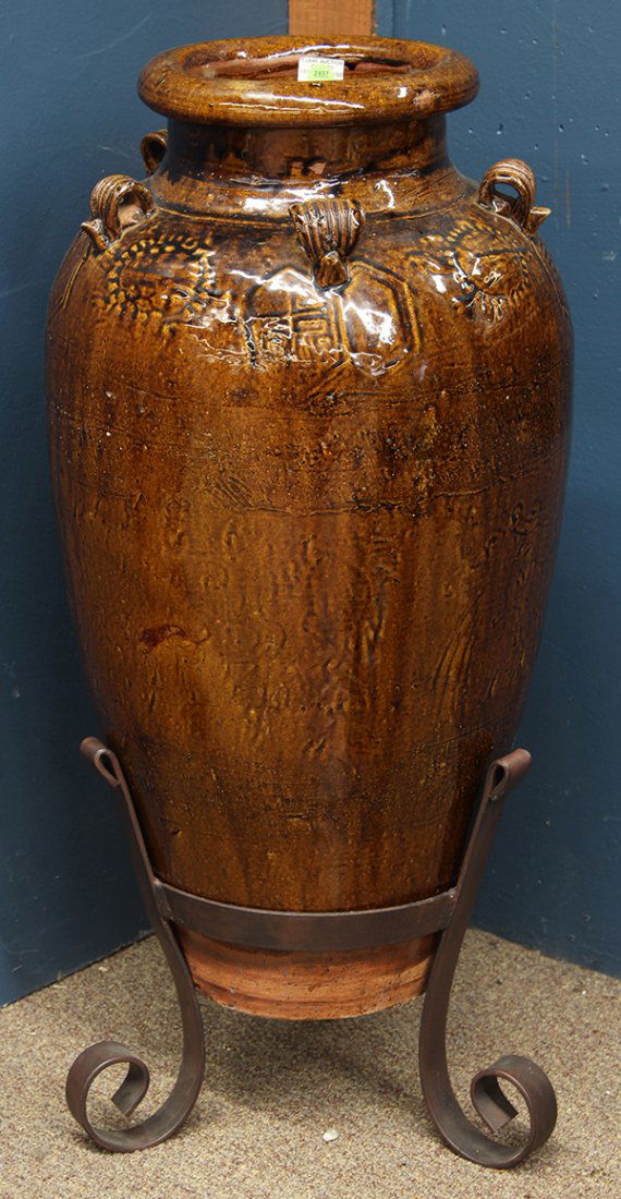 Large pottery urn: Large pottery urn, having applied handles and low relief decoration with brown high glaze and rising on a wrought iron stand, overall 32"h x 14"w
