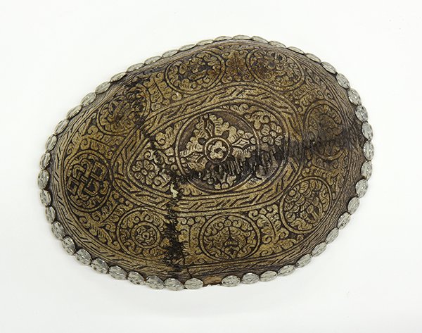 Sino-Tibetan Skull Cup: Himalayan skull cup, carved with the Eight Buddhist Treasures accented by foliage, the rim heightened with repousse skull appliques and a silver liner to the interior, 7"l. Provenance: From the estate