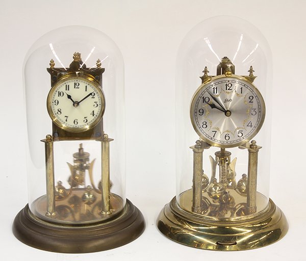 (lot of 2) German Anniversary clocks: (lot of 2) German Anniversary clocks, having glass domes with brass clocks, each with a white dial and Arabic numerals above a revolving pendulum, one marked Schatz, and each resting on a circular bra