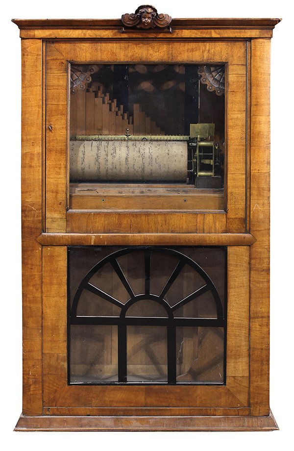 Biedermeier style cylinder organ having a pinned: Biedermeier style cylinder organ having a pinned cylinder and pipes, housed in an organ cabinet, having a putti carved crest, above a single door, surmounting a mullioned panel, 63"h x 40"w x 19"d
