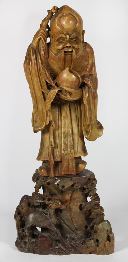 Chinese Soapstone Shoulao Sculpture: Chinese soapstone Shoulao sculpture, on a stand carved with rocks, lotus and deer, standing with a peach in his right hand and a staff in his left, 14"h