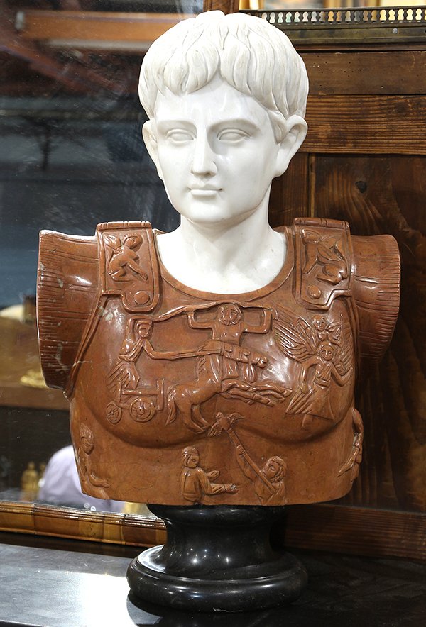 Classical style marble figural sculpture depicting a: Classical style marble figural sculpture depicting a Roman soldier, wearing armor decorated with a chariot, gazing outward with a distinguished expression, and rising on a black marble plinth, 22.5"h