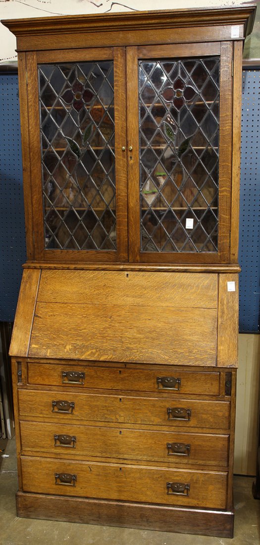 Secretary desk: Secretary desk, executed in quartersawn oak, having a stepped cornice above two leaded glass doors with polychrome floral designs, surmounting a slant front desk opening to a fitted interior, above fo
