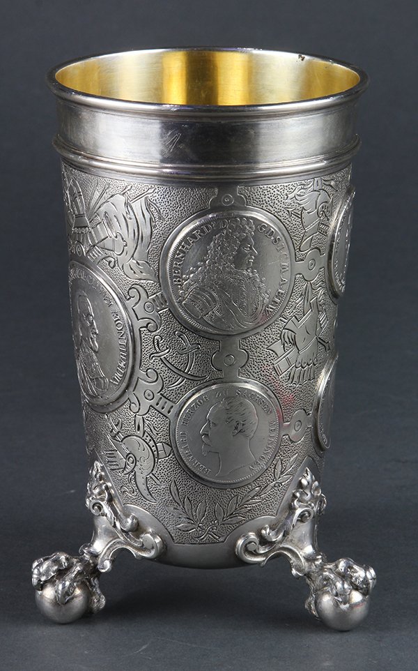 German: German .800 silver beaker, retailed by C.E. Keyser, Leipzig, the vessel having a molded band set over an intricately hammered ground detailed with ancient weaponry surrounding inset coins, and raised 