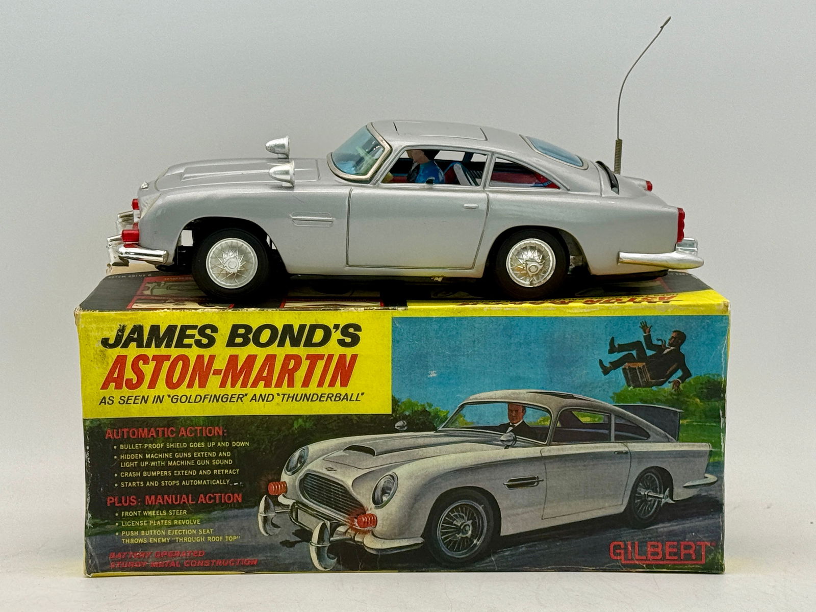 Gilbert James Bond Aston-Martin Toy Car with Reproduction Box: A silver/light grey toy James Bond Aston-Martin car with an antenna, presented on its reproduction colorful 1965 A.C. Gilbert Co. box labeled "JAMES BOND'S ASTON-MARTIN" and detailing "AUTOMATIC ACTIO