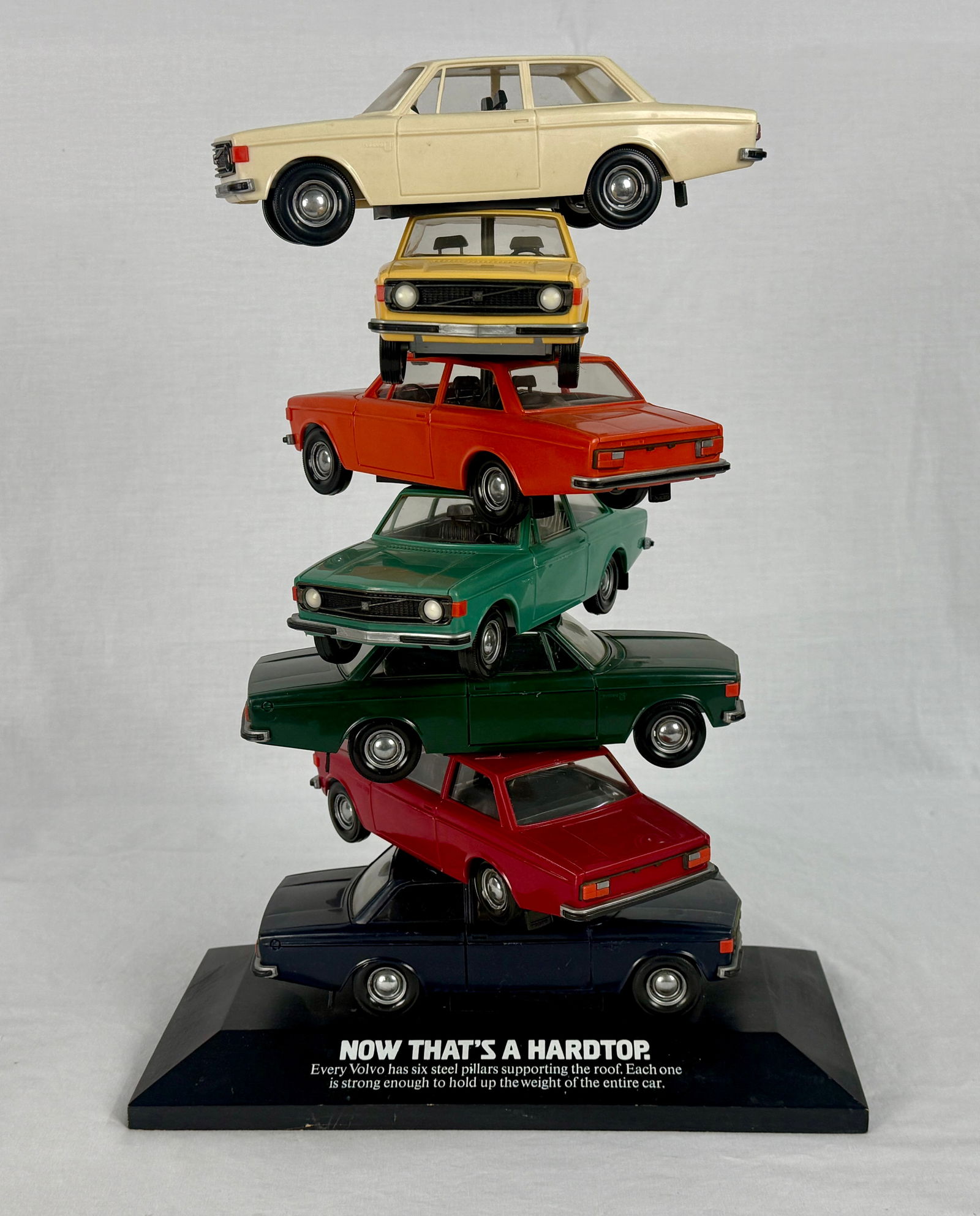 Vintage Volvo Promotional Model Car Display, 'NOW THAT'S A HARDTOP': Six toy/model cars, resembling Volvo sedans, in cream, yellow, orange, light green, dark green, and dark blue, are stacked on a black base labeled 'NOW THAT'S A HARDTOP.' and 'Every Volvo has a roof p