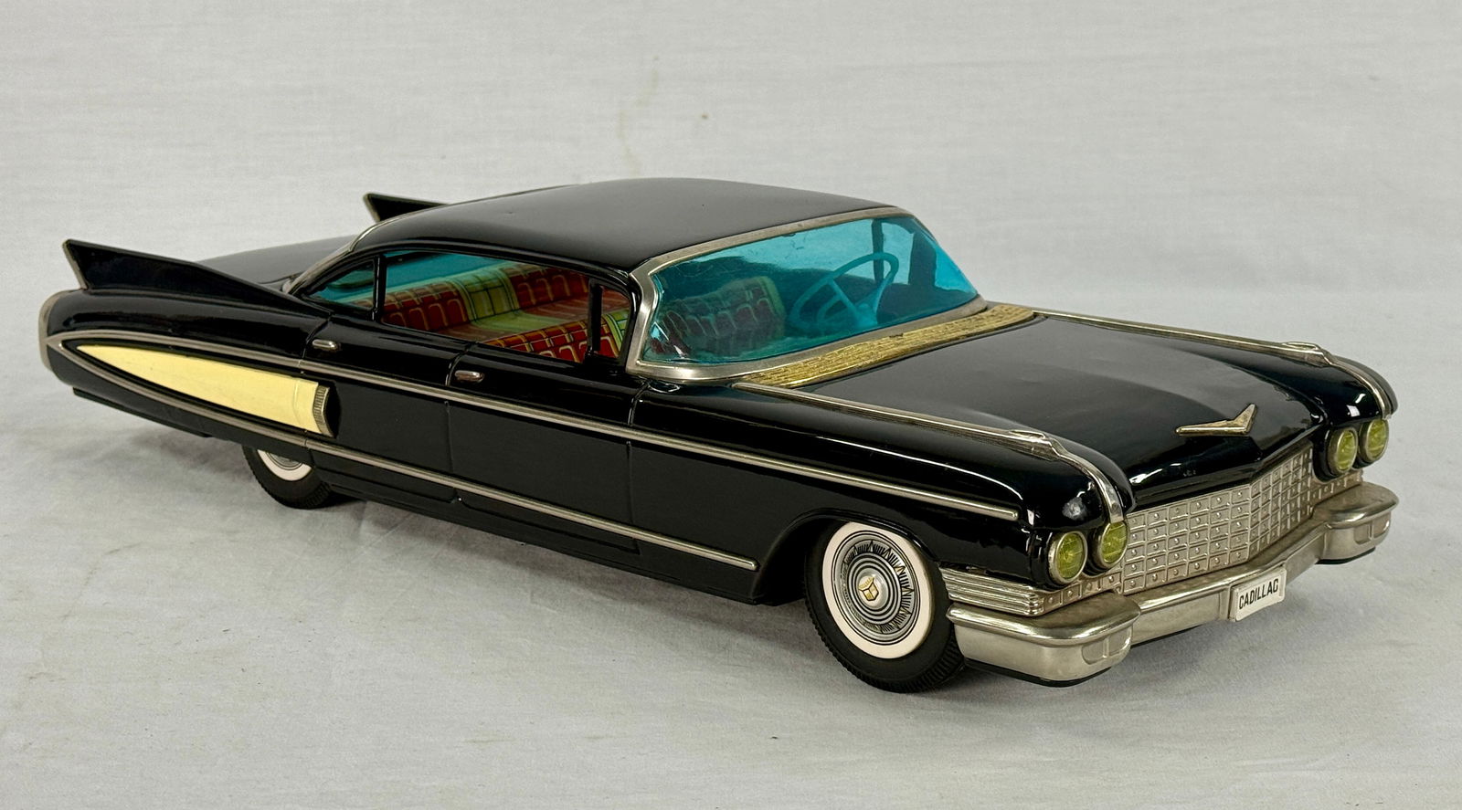 Yonezawa 1960 Cadillac Fleetwood Tin Friction Toy Car: A black Yonezawa 1960 Cadillac Fleetwood tin friction toy car with yellow and chrome side stripes, blue tinted windows, a red and yellow plaid interior, white wall tires, and "CADILLAC" on license pla