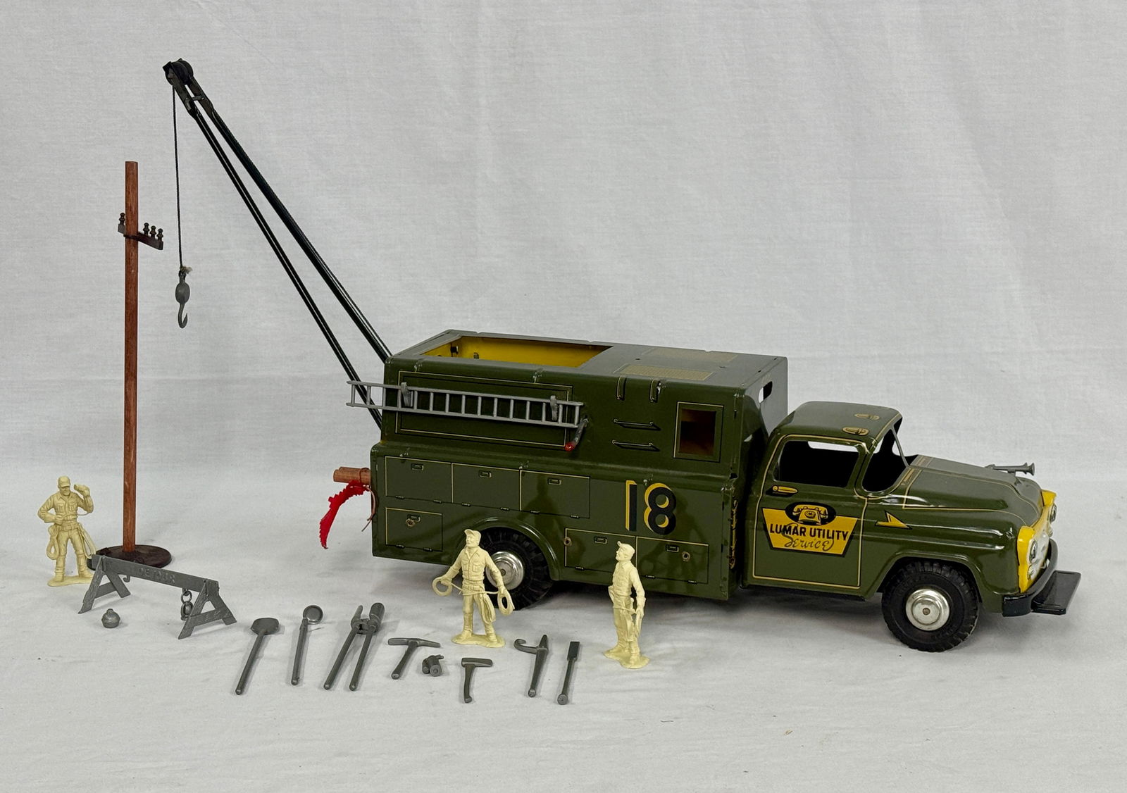 Marx Lumar Utility Service Truck Play Set: An olive green 20 inch Lumar Utility Co. Inc. N.Y.C. service truck with a black crane and a yellow '18' on its side, accompanied by a wooden utility pole, three beige plastic worker figures, and a gro