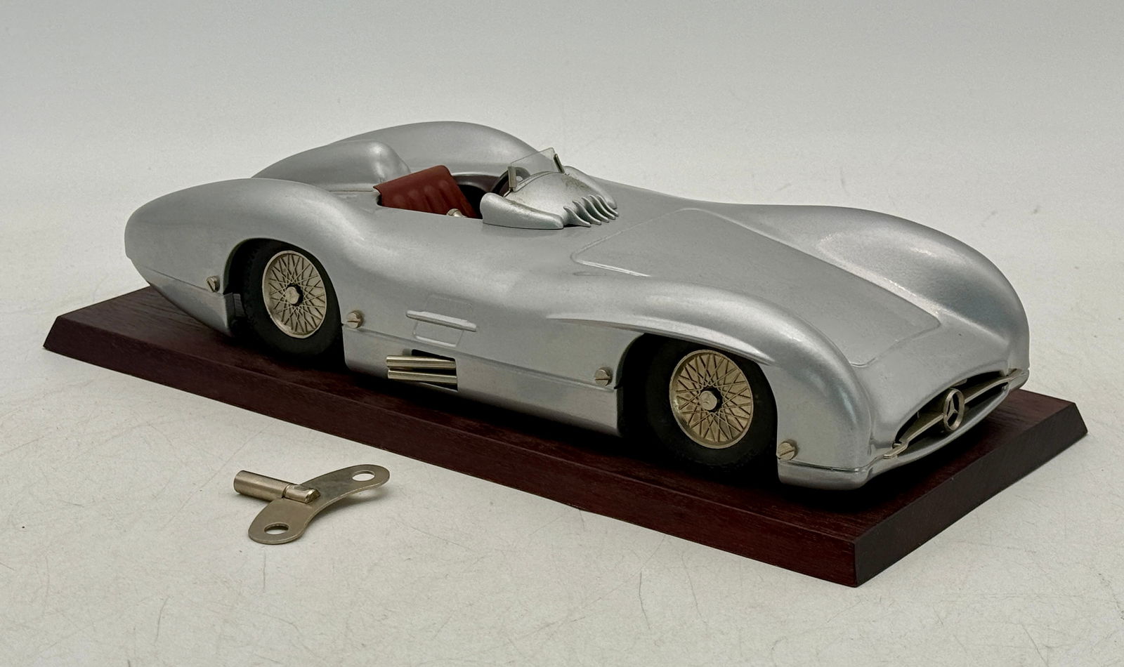 Marklin Mercedes-Benz Silver Race Car Model with Winding Key: A silver Marklin Mercedes-Benz race car model with brown interior, gold-colored spoked wheels, and star logo, displayed on a dark brown wooden base with a silver winding key C8.