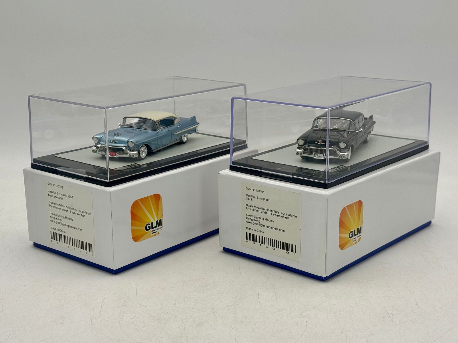 2 Great Lighting Models (GLM) 1/43 Scale 1957 Cadillac Model Vehicles: Two GLM 1/43 scale resin model vehicles including a blue metallic 1957 Cadillac Series 60 43100702 and a black Cadillac Brougham 43100101, each housed in a clear display case with original packaging.