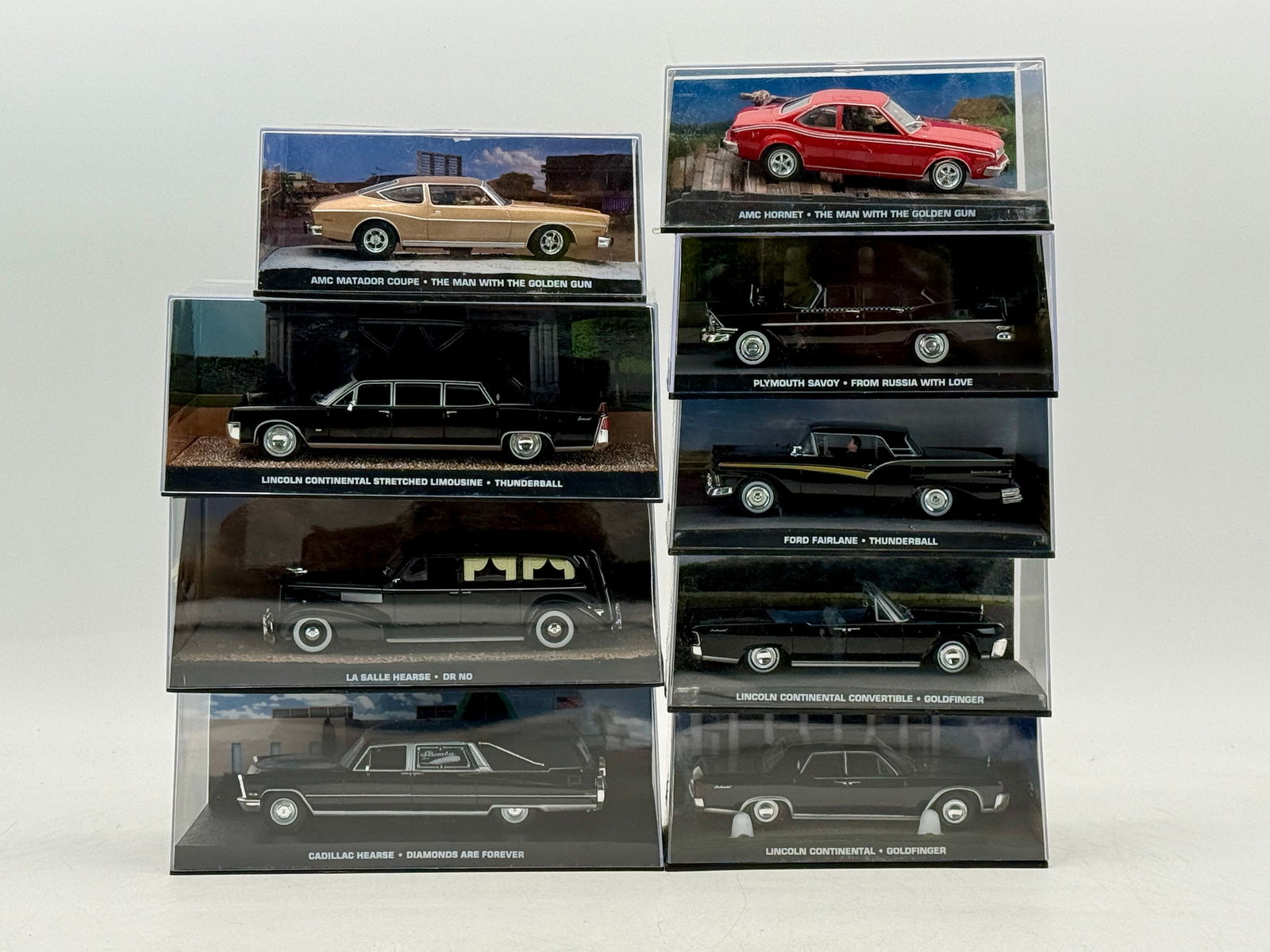 A Collection of 9 James Bond 007 Themed Model Cars: A collection of 9 die-cast/resin model cars featuring various makes including AMC, Lincoln, Plymouth, Ford, Cadillac, and La Salle, each housed in a clear display case with labels identifying their